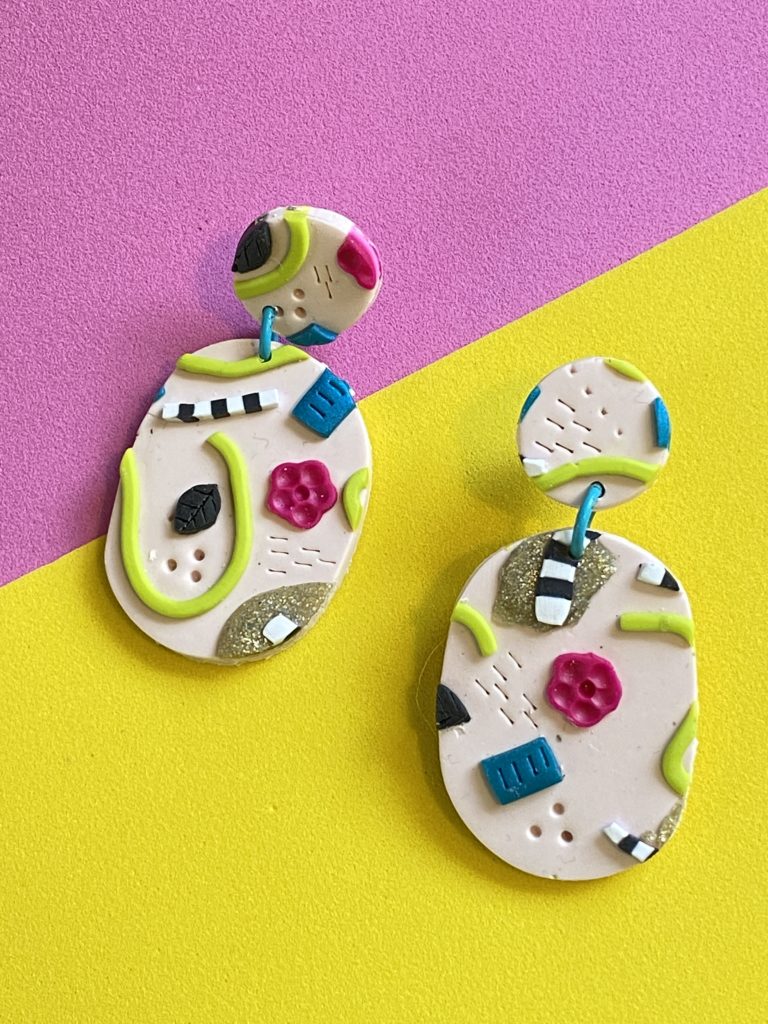 a pair of oval polymer clay dangle earrings with a pale pink background and bold colorful 3D designs on them.