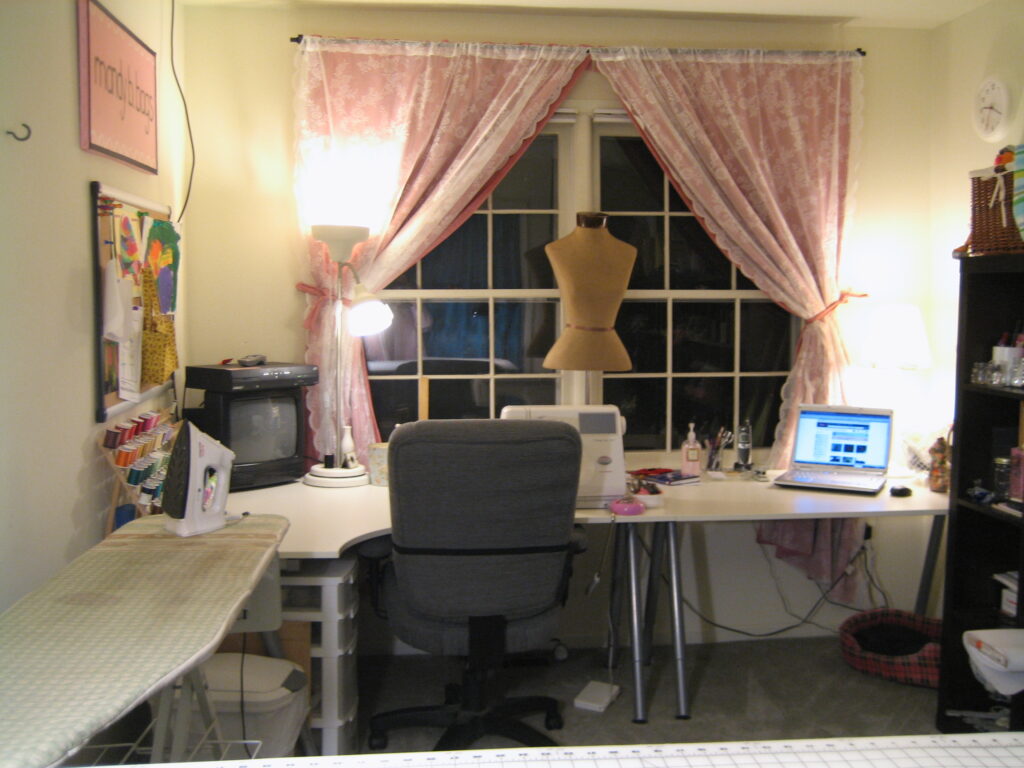 Image of a craft room depicting a long white table under a double window, including a chair, TV, sewing machine, computer, mannequin, light, and an ironing board.