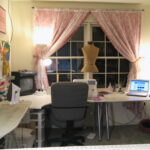 Image of a craft room depicting a long white table under a double window, including a chair, TV, sewing machine, computer, mannequin, light, and an ironing board.