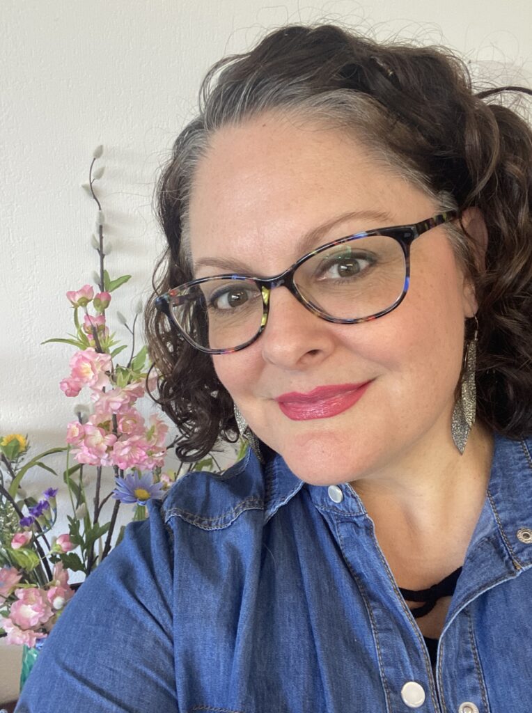 Image of Amanda Gendek, owner of One Cozy Nest. 
 She has curly shoulder-length brown hair with salt & pepper mixed in and is wearing glasses, a denim shirt, pink lipstick, posing next to colorful spring flowers.