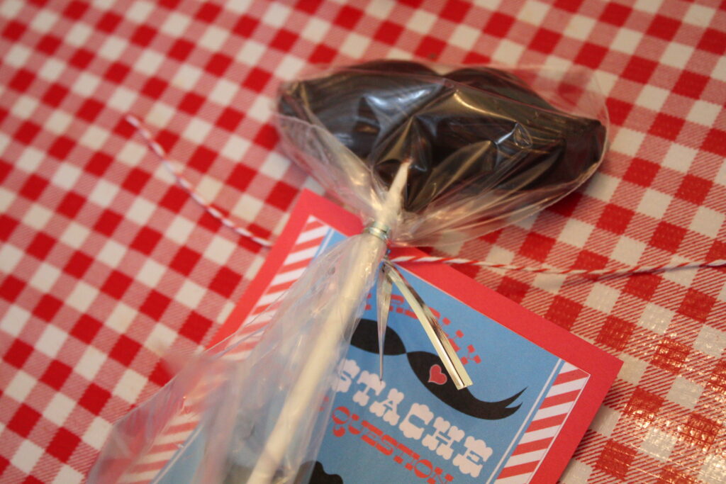 a chocolate mustache lollipop wrapped in plastic with a tag attached that reads "I mustache you a question".