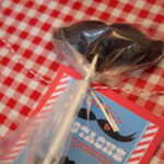 a chocolate mustache lollipop wrapped in plastic with a tag attached that reads "I mustache you a question".